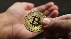 Ilustrasi tangan memegang smartphone menampilkan grafik Bitcoin, melambangkan kemudahan akses dan potensi investasi aset digital. Latar belakang menunjukkan koin Bitcoin yang berkilauan, menggambarkan nilai dan daya tariknya di pasar finansial modern.