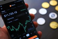 Seorang individu sedang serius memantau pergerakan grafik harga Bitcoin di layar gawai, mencerminkan aktivitas yang sering dilakukan oleh para investor kripto. Gambar ini menunjukkan bagaimana teknologi digital telah mengubah cara kita berinteraksi dengan aset investasi.