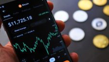 Seorang individu sedang serius memantau pergerakan grafik harga Bitcoin di layar gawai, mencerminkan aktivitas yang sering dilakukan oleh para investor kripto. Gambar ini menunjukkan bagaimana teknologi digital telah mengubah cara kita berinteraksi dengan aset investasi.