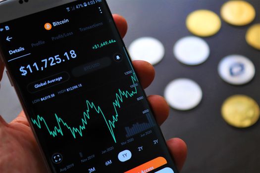 Seorang individu sedang serius memantau pergerakan grafik harga Bitcoin di layar gawai, mencerminkan aktivitas yang sering dilakukan oleh para investor kripto. Gambar ini menunjukkan bagaimana teknologi digital telah mengubah cara kita berinteraksi dengan aset investasi.